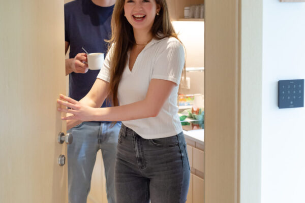 Das Bild zeigt zwei Personen, die in einem Türrahmen stehen. Die Dame im Vordergrund öffnet gerade die Tür, dahinter lacht die zweite Person hervor.