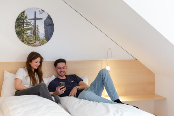 Das Bild zeigt zwei Personen, die auf einem Bett, die miteinander in ein Handy schauen. Über dem Bett hängt ein rundes Bild von einem Wald.