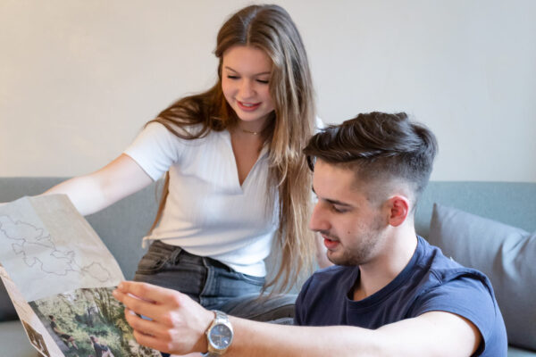 Das Bild zeigt zwei Personen, die auf einem Sofa sitzen und eine Karte oder Broschüre betrachten. An der Wand hinter ihnen befinden sich drei runde Bilder.